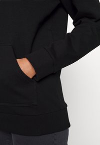 Zwarte sweatshirts met geribbelde manchetten en zoom. Voorzien van een zak aan de voorkant met een gladde textuur. Close-up van een hand in de zak.