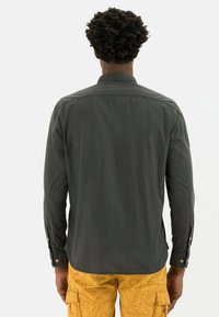 Chemise à manches longues vert foncé avec un col, présentant des poignets boutonnés et une texture lisse, assortie à un short cargo à motifs jaunes.