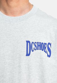 Heather grey sweatshirt with a ribbed crew neckline and a bold blue "DCSHOES" graphic on the front. Soft fabric texture, casual design.