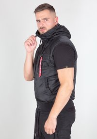 Melna bez piedurknēm puffer veste ar kapuci, ar rāvējslēdzēju priekšpusē, aukliņu, divām priekšējām kabatām un sarkanas markētas auduma etiķetes detaļu.