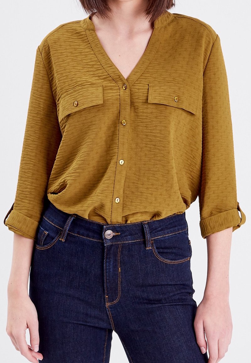 Chemise couleur moutarde avec un col en V, deux poches avant et des boutons de fermeture. Elle a un tissu texturé et des manches retroussées, associée à un jean foncé.
