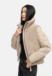 Beige jack met gemengde materialen met een pluizige bont voorkant en gestikte mouwen, hoge kraag en ritssluiting. Accenten van ribbels bij de manchetten en onderrand.