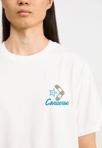 Balta 100% medvilnės marškinėlių su mėlyna ir oranžine grafika, įskaitant žvaigždę ir pleistro dizainą, su tekstenis "Converse" po jais.