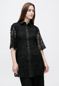 Μαύρο δαντελένιο πουκάμισο με κουμπιά στο μπροστινό μέρος, κοντά μανίκια και γιακά. Πολύπλοκα λουλουδάτα σχέδια διακοσμούν το ύφασμα, με μια στερεή μαύρη λωρίδα με κουμπιά.