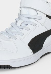 Λευκό ψηλό αθλητικό παπούτσι με μαύρη πλαϊνή ρίγα, λευκά κορδόνια και λουράκι με βέλκρο πάνω από τον αστράγαλο.