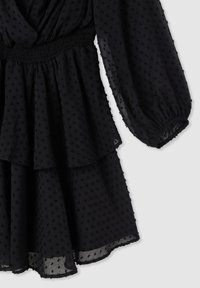 Czarna sukienka w kropki, wykonana z przezroczystego materiału. Posiada gumiak w talii, wielowarstwowe falbany oraz długie rękawy z elastycznymi mankietami.