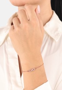 Pulsera de oro rosa con un símbolo de infinito adornado con pequeñas piedras transparentes, llevada en una muñeca delicada contra una camisa de color claro.