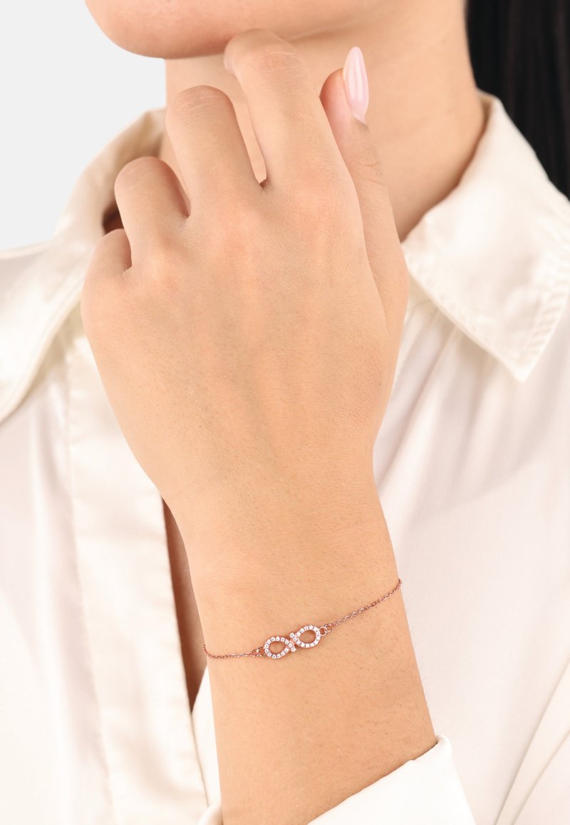 Pulsera de oro rosa con un símbolo de infinito adornado con pequeñas piedras transparentes, llevada en una muñeca delicada contra una camisa de color claro.