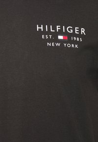 Czarna tkanina z białym napisem "HILFIGER EST. 1985 NOWY JORK" oraz małym prostokątnym logo w kolorach białym, czerwonym i granatowym.