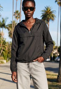 Vrouw in een zwarte windjack en zonnebril staat buiten met palmboomachtergrond op een zonnige dag.