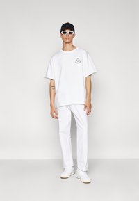 Tricou alb supradimensionat cu detalii grafice, pantaloni albi asortați, adidași albi și o șapcă neagră. Textură netedă, design casual.