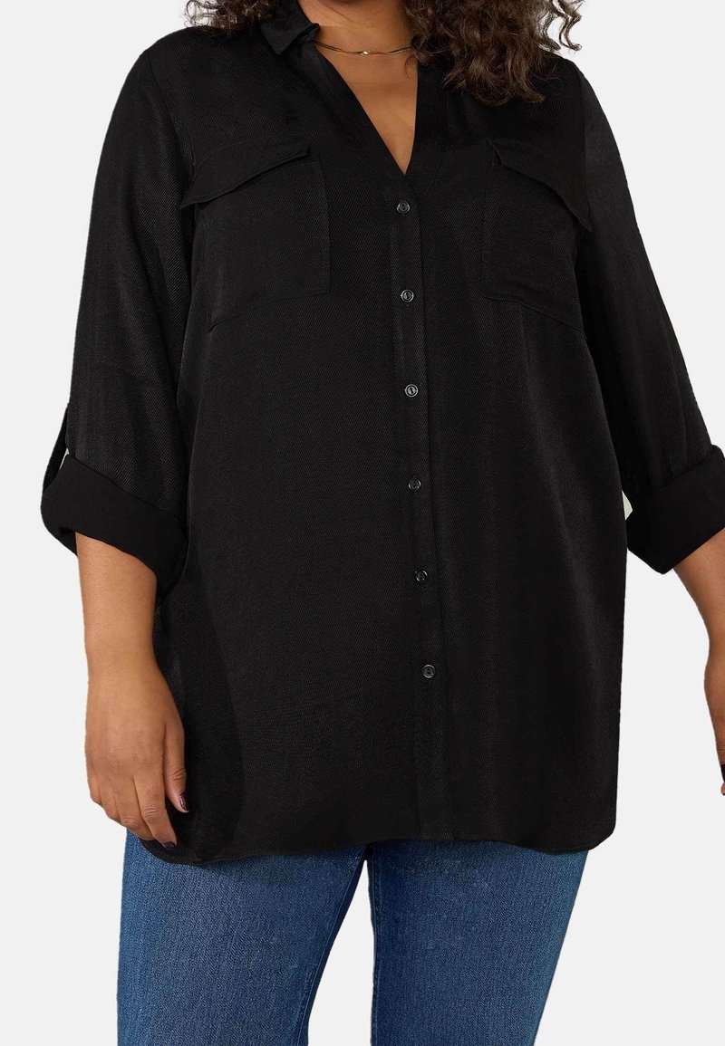 Camicia nera con colletto, due taschini sul petto, maniche arrotolate e tessuto strutturato. Abbinata a jeans blu.