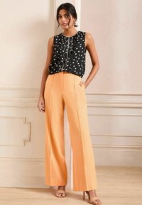 Musthave musta ilma varrukatega topp valgete täppidega ja dekoratiivne eesmine paneel, millele on lisatud kõrge vöökoht ja laiad oranžid püksid.