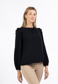 Donna che indossa una camicetta nera a maniche lunghe con dettagli a volant e pantaloni beige, posando su uno sfondo bianco semplice.
