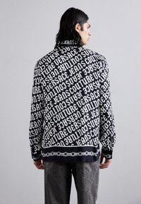 Μακριά μπλούζα με μαύρο χρώμα και έντονη λευκή εκτύπωση που περιλαμβάνει επαναλαμβανόμενο κείμενο. Λείος ύφασμα, χαλαρή εφαρμογή και αντιθετικό σχέδιο αλυσίδας στο τελείωμα.