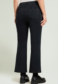 Zwarte uitlopende jeans met een hoge taille, voorzien van twee achterzakken en een gladde textuur. Gepaard met zwarte instappers en sokken.