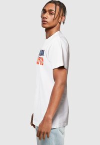 Tricou din bumbac alb cu text imprimat în nuanțe de albastru și portocaliu. Design clasic cu guler rotund și mâneci scurte, prezentat pe un model în profil.