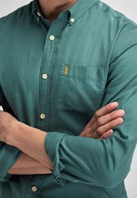 Camicetta verde con bottoni e taschino, caratterizzata da un piccolo ricamo di un'aragosta dorata. Il tessuto è testurizzato e morbido, con dettagli classici sui polsini.