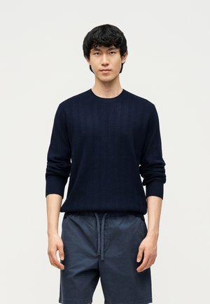 Un jeune homme se tient face à nous, portant un pull à manches longues bleu marine foncé et un short assorti bleu marine avec une taille à cordon.