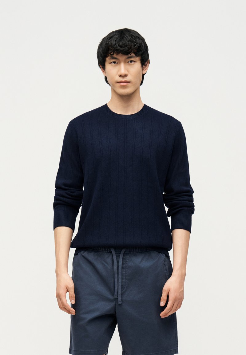 Un jeune homme se tient face à nous, portant un pull à manches longues bleu marine foncé et un short assorti bleu marine avec une taille à cordon.