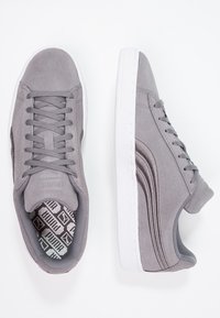 Harmaat mokka-sneakerit, joissa on valkoiset kumipohjat, litteät nauhat ja kuvioitu sisäpohja. Suunnittelussa on hienovaraisia sivuaksentteja ja teksturoitu pinta.