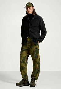 Νεαρός άνδρας φορώντας μαύρο μπουφάν, καμουφλάζ παντελόνι cargo, μαύρα αθλητικά παπούτσια και ελιά-πράσινο καπέλο, στέκεται με τα χέρια στις τσέπες.