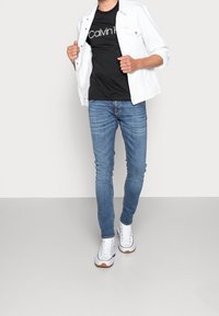Bílá denimová bunda přes černé tričko s logem, spárovaná s světle modrými skinny džíny a bílými teniskami s hnědými akcenty.