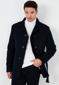 Tengerészkék gyapjú keverék kabát dupla gombos záródással, vállpántokkal és derékig érő övvel, fehér garbó alatt.