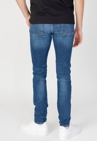 Jeans in denim blu con una vestibilità slim, caratterizzati da tasche posteriori con dettagli di cucitura e una texture consumata, abbinati a sneakers bianche.