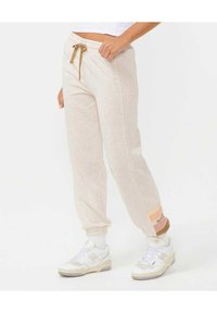 Beige sweatpants med dragsko i midjan, avsmalnande manchetter och ett rosa accentband nertill med texten "nid". Mjuk, texturerad material.