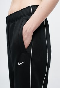 Pantaloni sportivi negri cu margini albe pe laterale, talie elastică și buzunare laterale. Prezintă un mic logo alb Nike pe șoldul stâng.