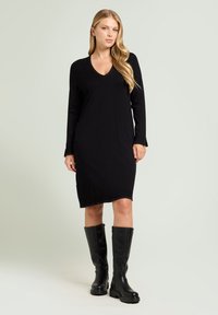 Robe noire en maille à manches longues avec un décolleté en V, arrivant à mi-cuisse, portée avec des bottes noires hautes jusqu'au genou. Texture lisse et design simple.