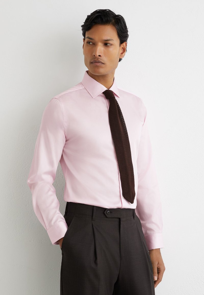 Homme portant une chemise rose clair et une cravate tricotée brun foncé avec un pantalon foncé, debout contre un mur blanc, mains dans les poches.