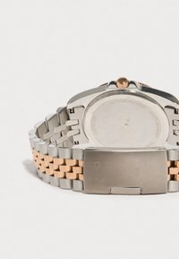 Kaksi sävyinen metallinen rannekello, jossa on "LIU JO"-kaiverruksella varustettu solki, näyttäen takakannen yksityiskohtia ja kruunua valkoista taustaa vasten.