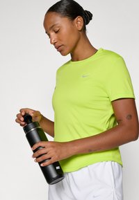 Jasnozielona koszulka sportowa z krótkim rękawem i logo Nike. Czarna butelka na wodę z srebrnymi akcentami trzymana w jednej ręce. Białe szorty.