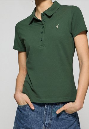 Grøn polo skjorte med ensfarvet design, korte ærmer, krave og fem knapper. Har et lille, cremefarvet logo på venstre bryst.