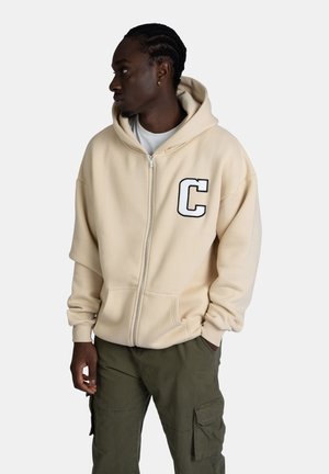 Beige zip-up hoodie with a large black 'C' on the chest. Features a drawstring hood and kangaroo pocket. Worn with olive green cargo pants.