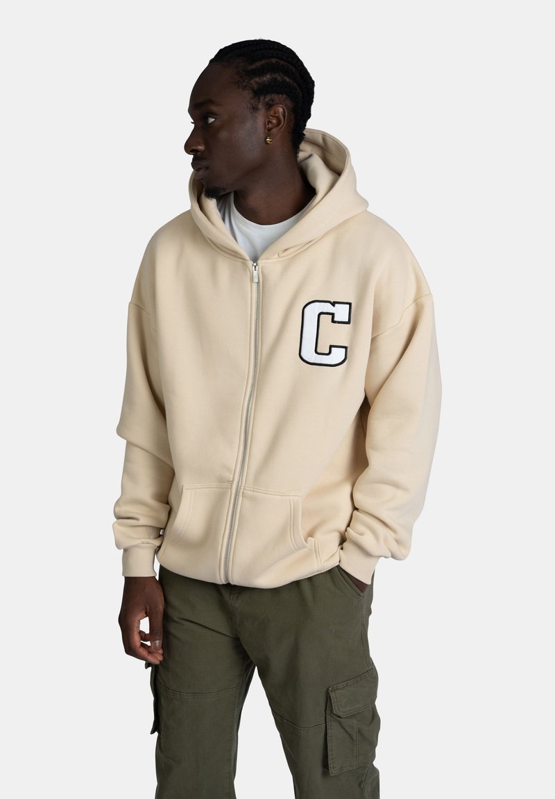 Felpa con zip beige con una grande 'C' nera sul petto. Presenta un cappuccio con lacci e tasca a marsupio. Indossata con pantaloni cargo verde oliva.