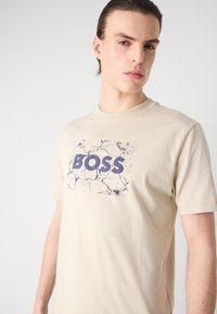 Béžové bavlněné tričko s kulatým výstřihem a krátkými rukávy. Na pozadí s mramorovým vzorem se nachází modré logo "BOSS".