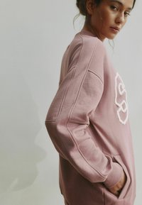 Roosa lõdva lõikega dressipluus, millel on tekstuuriga valge logo, soonikkoes mansetid ja küljetaskud. Kangas näib pehme ja mugav.