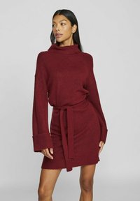 Robe tricotée bordeaux avec un col haut, des manches longues et une ceinture nouée à la taille. Présente des poignets côtelés et une texture douce.
