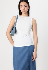 Weißes ärmelloses Oberteil mit Rundhalsausschnitt, kombiniert mit einem blauen Jeansrock mit einem Schlitz vorne, gehalten mit einer kleinen blauen Schultertasche.