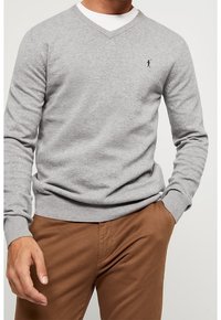 Pull gris en maille avec un col en V et des poignets côtelés, ornée d'un petit logo noir brodé sur la poitrine. Associé à un pantalon marron.