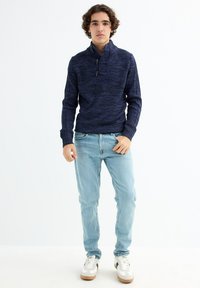 Suéter de punto azul marino con un cuello alto y detalles de botones, combinado con jeans azul claro y zapatillas blancas con detalles en marrón.