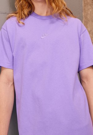 Osoba nosząca lawendowy t-shirt Nike z krótkimi rękawami i wyszywanym logo na środku klatki piersiowej.