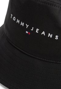 Μαύρο καπέλο τύπου bucket κατασκευασμένο από ύφασμα με λεία υφή. Διαθέτει λευκό κεντημένο λογότυπο "TOMMYJEANS" και ένα μικρό τρίχρωμο έμβλημα.