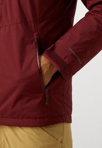 Red waterproof jacket with a textured finish. Features zippered side pocket and adjustable sleeve cuff. Logo displayed on the sleeve.