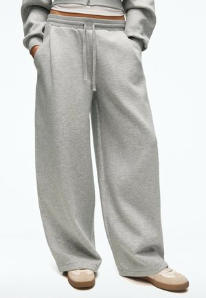 Personne portant un pantalon de jogging gris ample avec cordon de serrage et les mains dans les poches, associé à des baskets beiges et blanches.