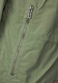 Tissu vert avec fermeture éclair argentée verticale et coutures doubles sur un vêtement, gros plan montrant les tirettes de fermeture-éclair étiquetées "CECIL".