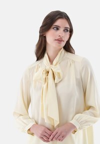 Gelbe Seidenbluse mit einer Schleifendetail, langen Ärmeln mit Knopfmanschetten und gerafften Schulternähten für eine fließende Silhouette.